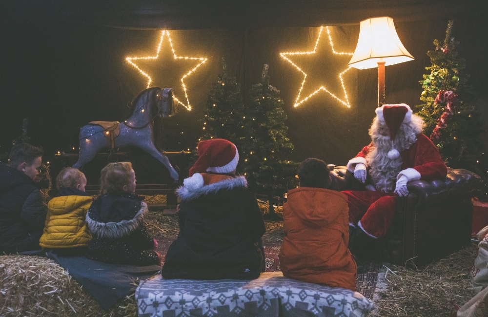 Santa in a barn with fairy lights and children gathered around