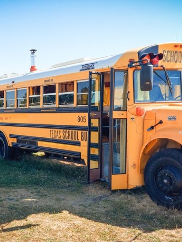Big yellow American school bus in a field next to wooden bench and umbrella