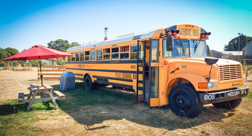 Big yellow American school bus in a field next to wooden bench and umbrella