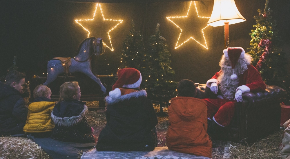 Santa in a barn with fairy lights and children gathered around